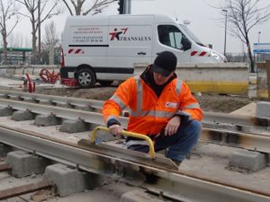 Un ouvrier en veste orange haute visibilité s'accroupit sur des rails de tramway en chantier, avec une camionnette blanche en arrière-plan.