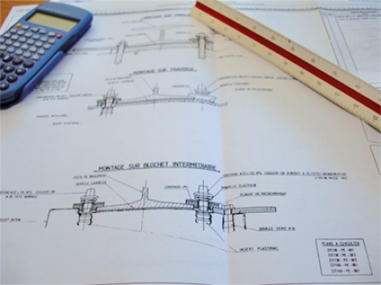 Des plans techniques de montage en français posés sur une table, avec une calculatrice bleue et une règle d'échelle rouge et blanche.