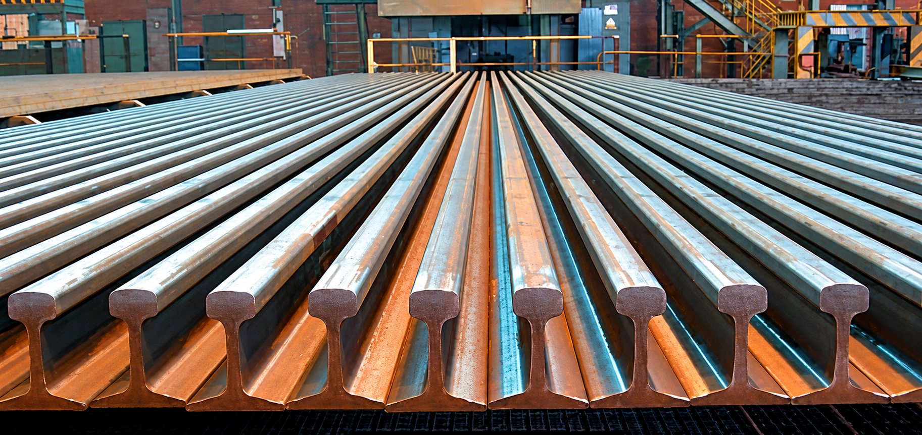 De nombreux rails d'acier posés côte à côte en rangées dans un atelier industriel, vus depuis l'extrémité.