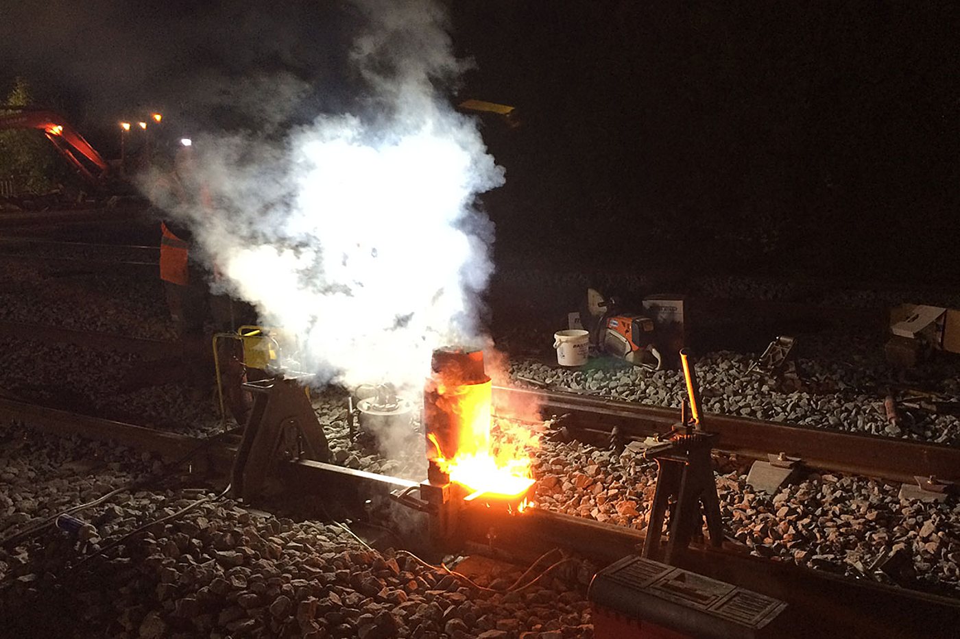 De nuit sur une voie ferrée, une soudure aluminothermique de rail dégage de la fumée et des flammes orangées, avec des outils posés sur le ballast.